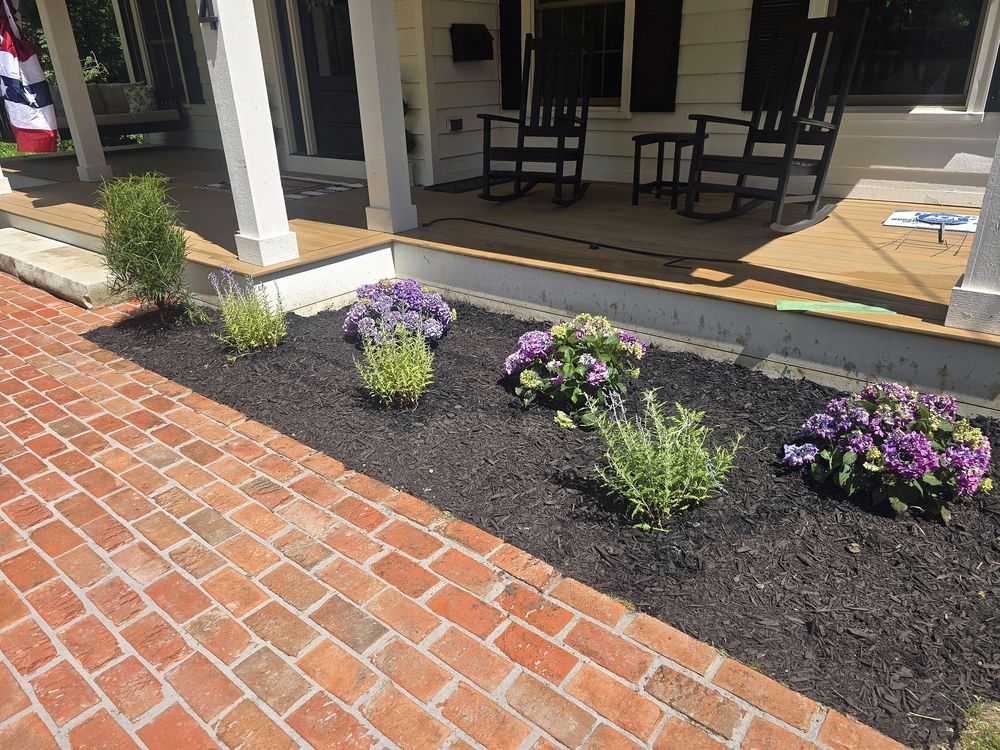 Red brick walkway beside black mulch beds and a white farmhouse porch