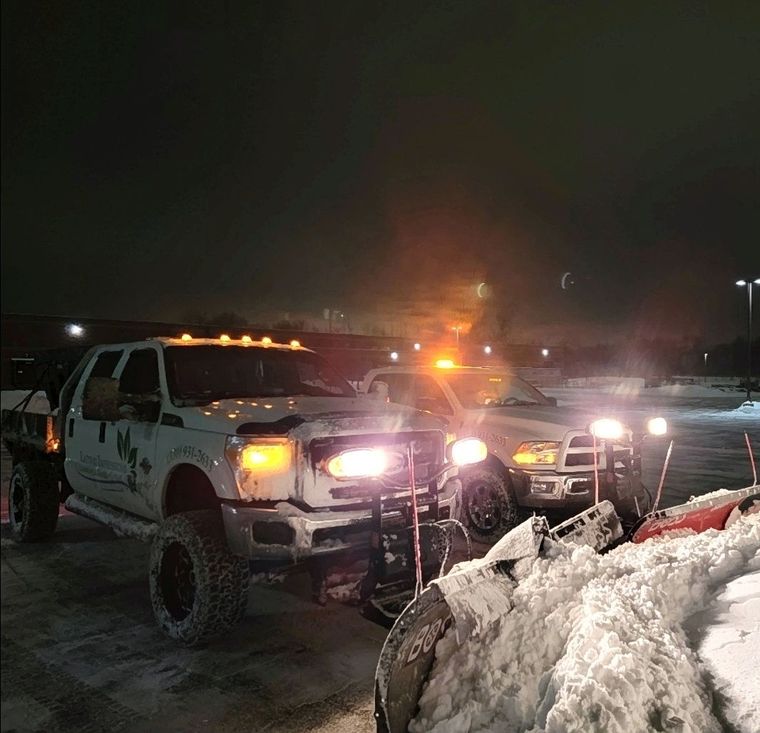 Two branded plow trucks working a commercial lot at night under amber beacons